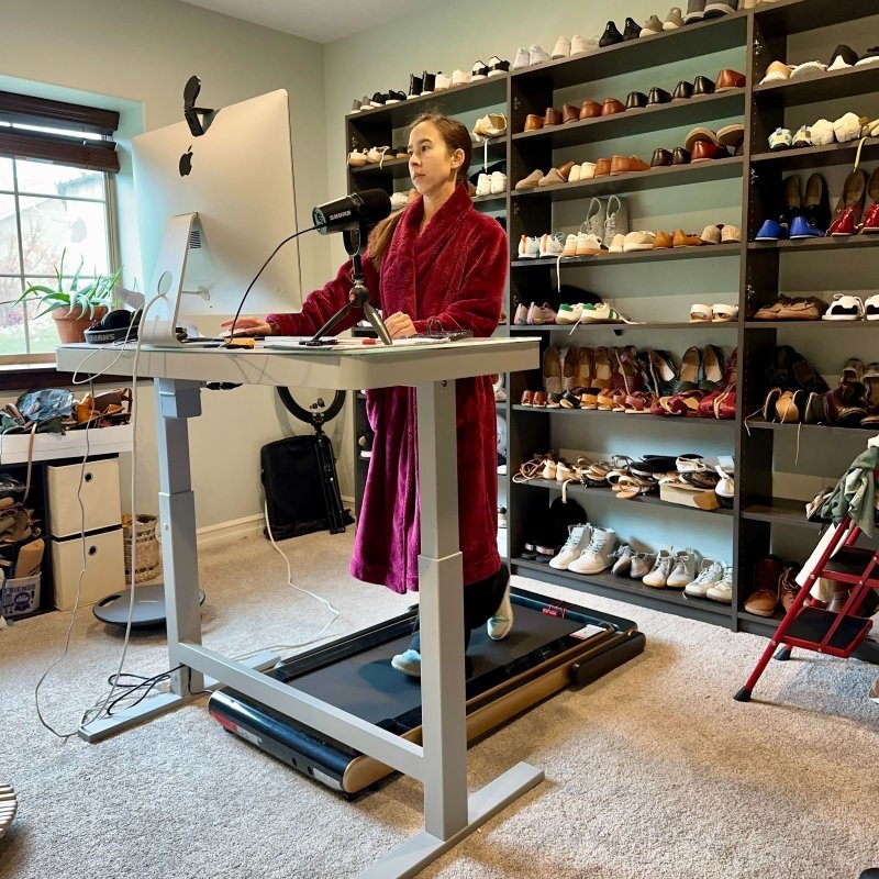 A woman in a fuzzy red robe walking on a walking pad at a standing desk in front of a wall of shelves filled with barefoot shoes
