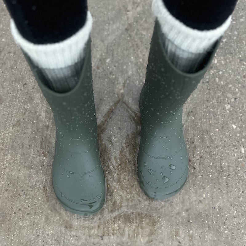 A top down view of a pair of Wildling Shoes Ranidae rubber rain boots standing in a puddle, with wool socks peeking out of the top.