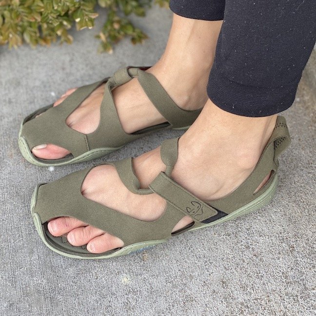 A side view of a pair of feet standing on concrete wearing Wildling Shoes Forest Feather barefoot summer sandals in green microfiber