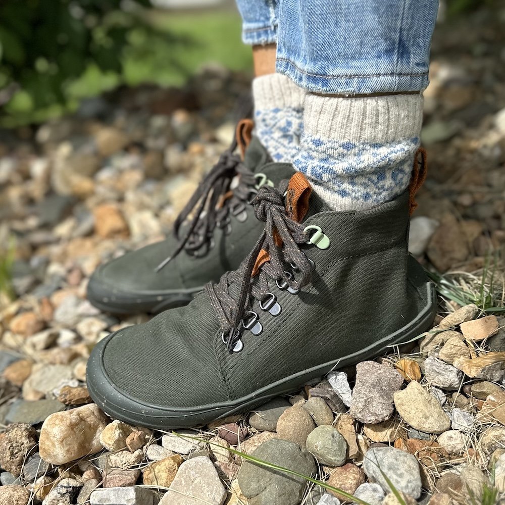 A close up side view of a pair of feet standing on rocks outdoors wearing wool socks and Wildling barefoot shoes Tejo fall boots in olive green vegan material
