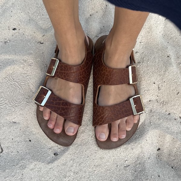 A person's feet in Wildstride Buffalo Leather barefoot Birkenstock style sandals sitting on a sandy beach. The chunky straps are textured and set off with gold buckles.