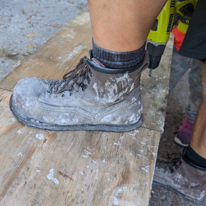A man wearing Carets Determination Safety Steel Toe Boots with 1 foot on a piece of plywood to steady it while working. The boots are well worn and visibly splattered with a chalky white/gray substance.
