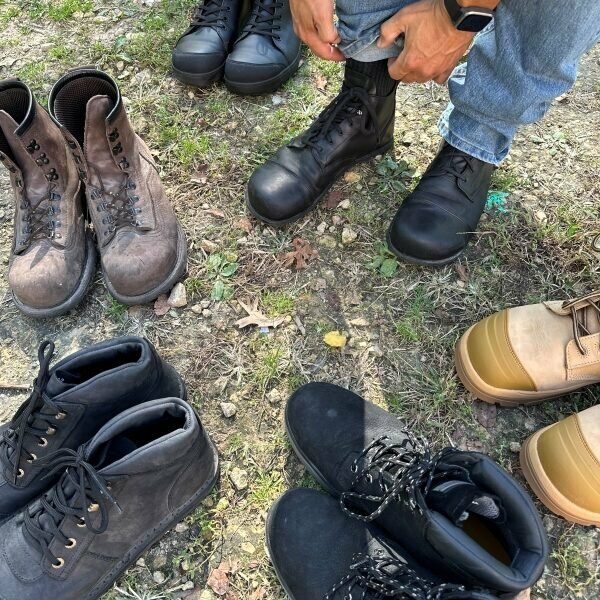 A man putting on a pair of wide toe box steel toe safety boots from Gaucho Ninja. He is surrounded by other wide toe box barefoot friendly steel toe options from Carets, DeWalt, Jim Green, Birkenstock, and Wide Load.