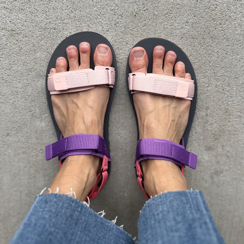 A top down view of a pair of feet standing on pavement wearing colorful Xero Shoes D Trail barefoot all purpose sandals