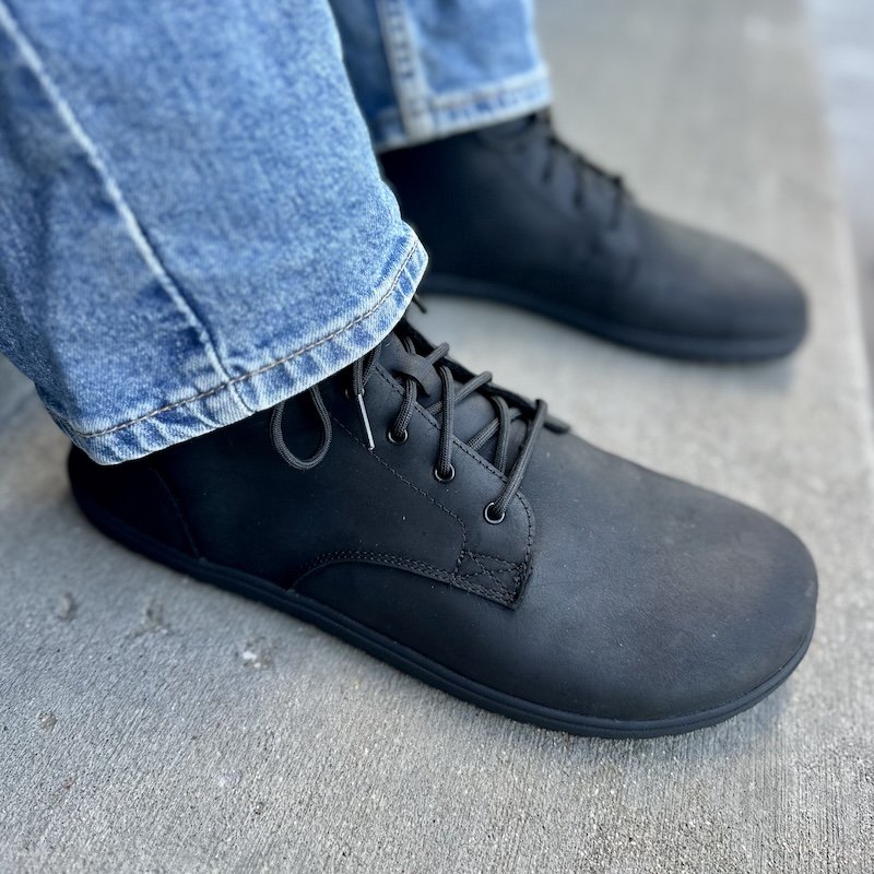 Close up side angled view of a pair of Xero Shoes Denver II Men's Barefoot leather boots in black with a pair of mid-tone denim sitting at the ankle.