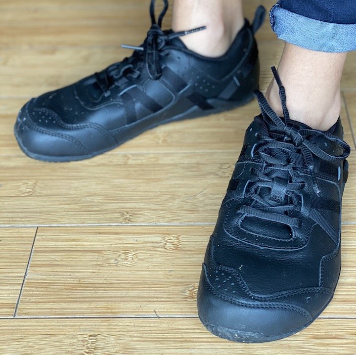A front view of a pair of feet standing on a wood floor wearing the Xero Shoes Prio All-Day barefoot sneaker made for the service industry with a non slip sole in black leather