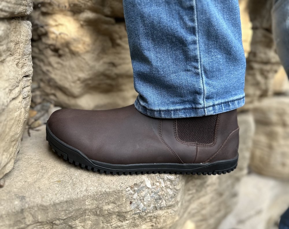 Side view of a man's foot in Xero Shoes Ridgeway chelsea boot stepping on part of a stone wall. The black zero drop outsole is visible and the color matched elastic on the side is peeking out from under medium wash jeans.