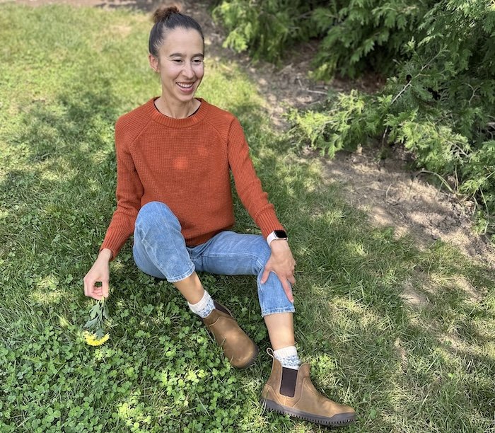A woman sitting on grass wearing a burnt orange sweater, jeans, and Xero Ridgeway Chelsea rugged barefoot boots with a wide toe box in brown leather