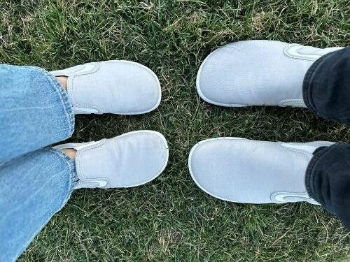 Top down view of 2 people wearing the same casual gray canvas slip on from Xero Shoes. The wide toe boxed shoes are toe to toe and the bottom pair of larger than the top