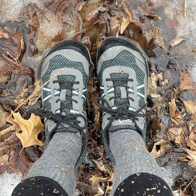 A top down view of a pair of feet standing on dead leaves, ice, and muddy water wearing the Xero Shoes terraflex barefoot hiking shoe in green and black