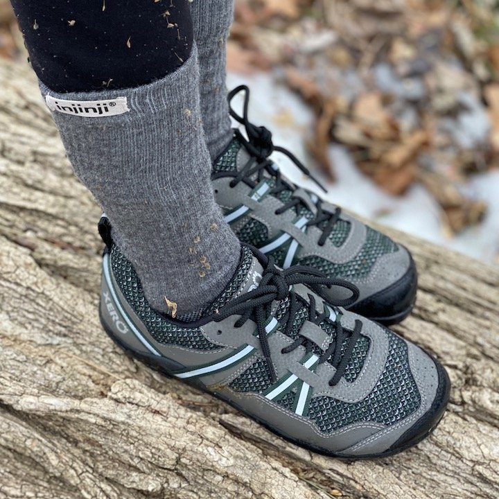 A tside view of a pair of feet standing on a log outside wearing the Xero Shoes terraflex barefoot hiking shoe in green and black