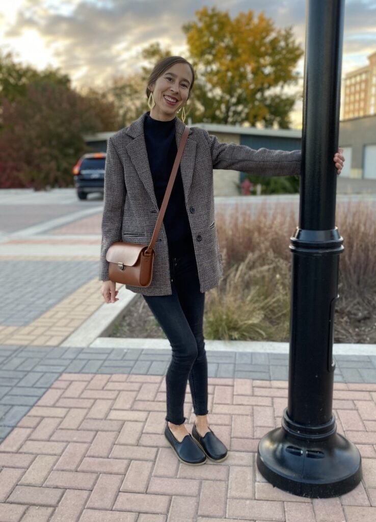 A woman wearing a plaid blazed and black pants smiling at the camera wearing Designed by Anya black Yasemin barefoot loafers