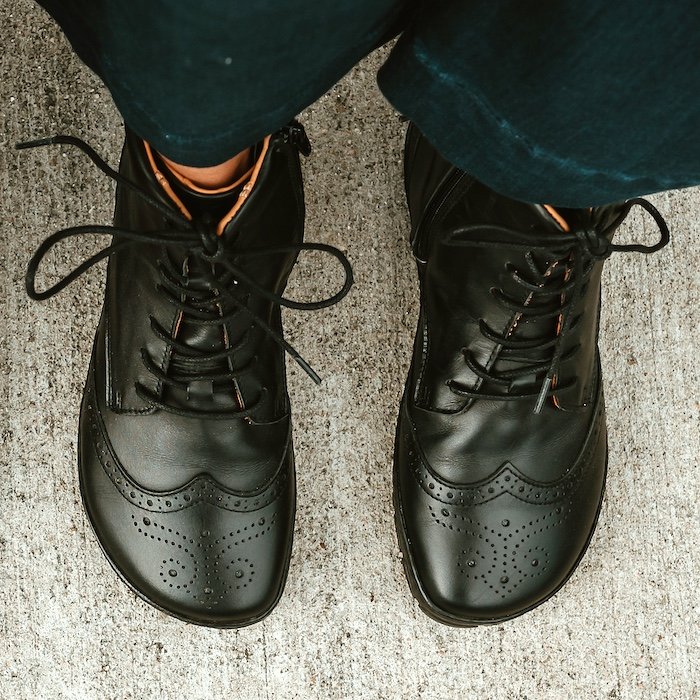 A top down view of a person's feet wearing black leather Zaqq Quintic brogue detailed wide toe box barefoot combat boots
