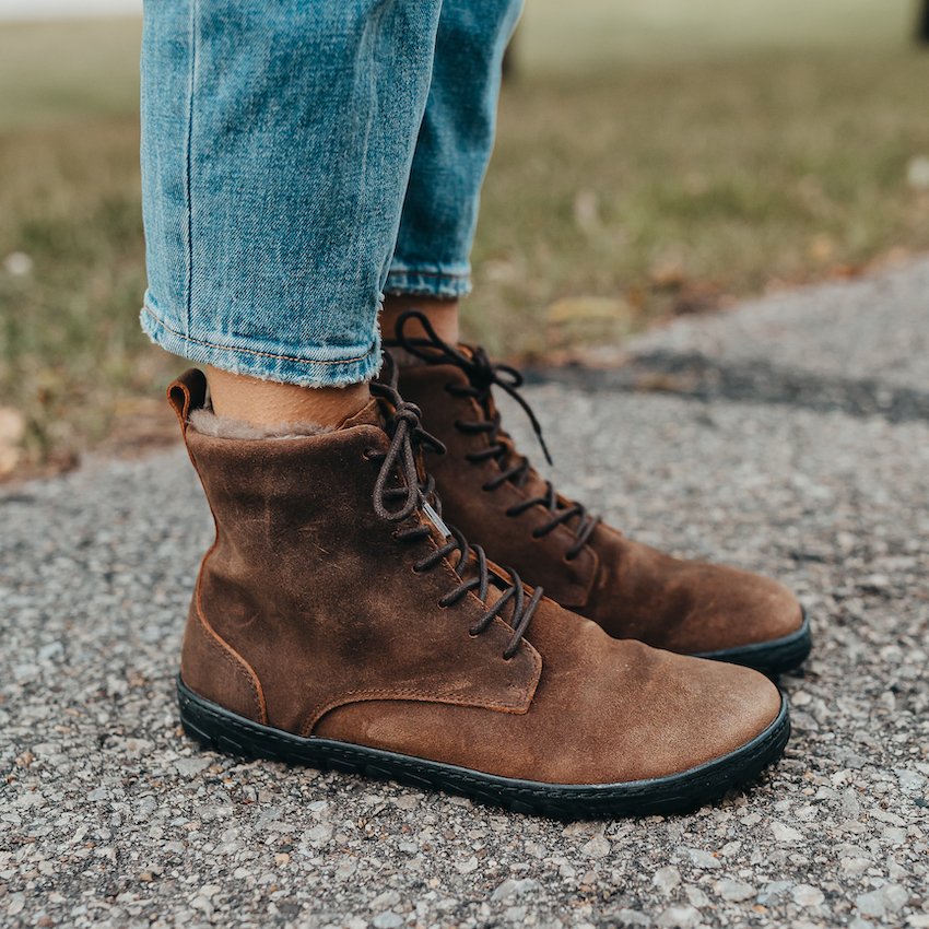 a close up side view of a person's feet wearing jeans and brown barefoot winter boots called the Quintic that are made of leather, fur lined, and waterproof.
