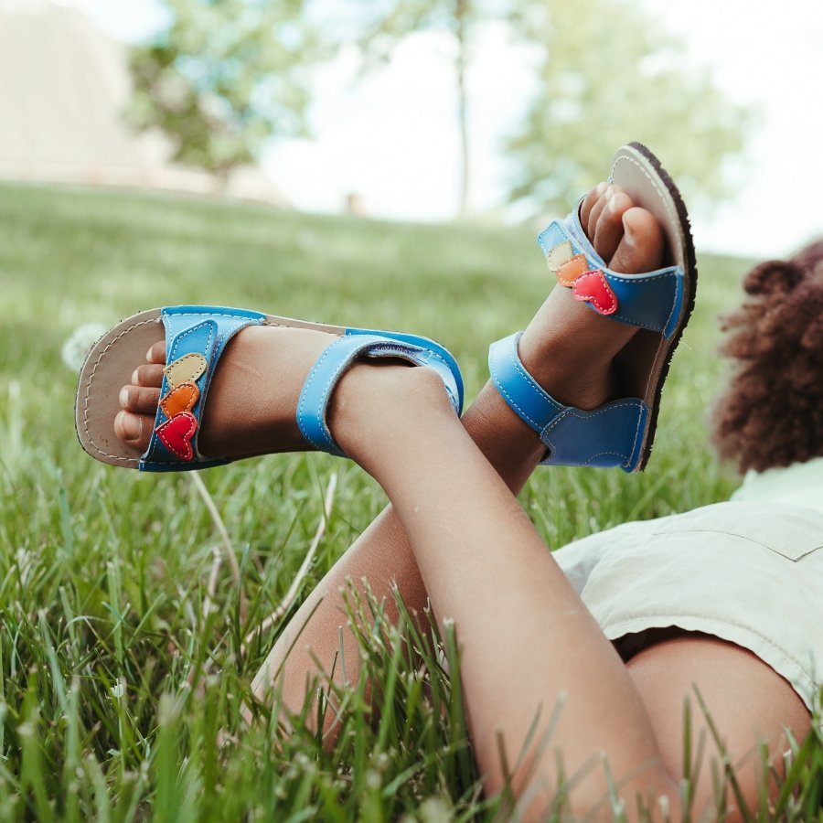 A photo from the back of a young child laying in the grass with their legs crossed. They have a pair of Zeazoo Ariel barefoot sandals on their feet featuring a wide toe box and high quality leather straps