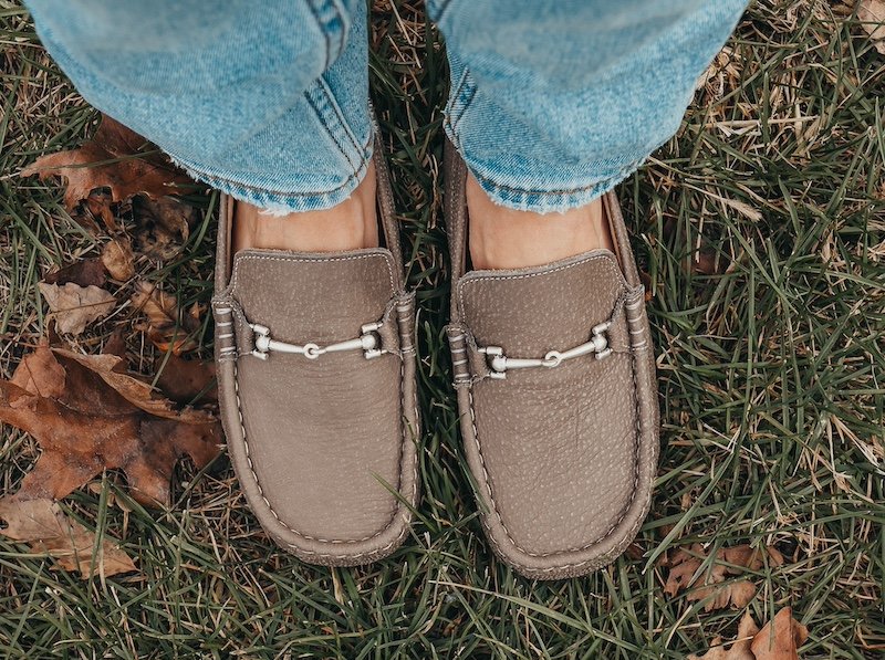 A top down view of a pair of feet standing in grass wearing Zeazoo Cheetah custom barefoot loafers in taupe with a metal buckle and a wide toe box